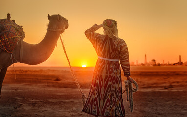 Woman in traditional national clothing leads camel through desert towards ancient city of Khiva at sunset. - obrazy, fototapety, plakaty