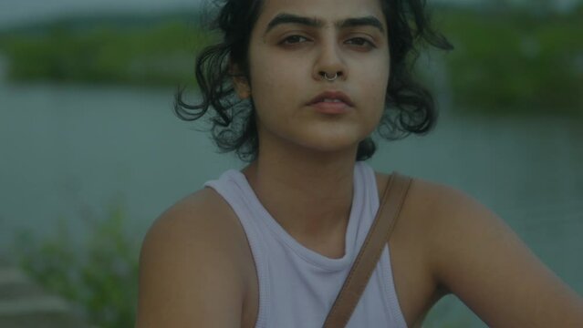 In slow motion, a captivating young Indian woman stands beside a tranquil river. She gracefully turns around and  faces the camera. Looking at the camera exuding an air of serene confidence and charm.