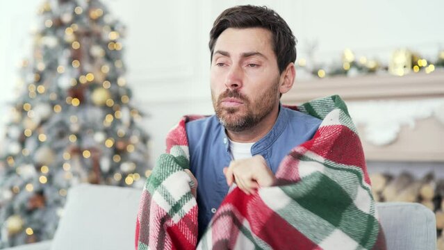 Upset bearded man covered with a blanket froze sitting on sofa in living room at home during winter New Year Christmas Xmas holidays. Male cold indoors. Guy feels discomfort due to low temperature