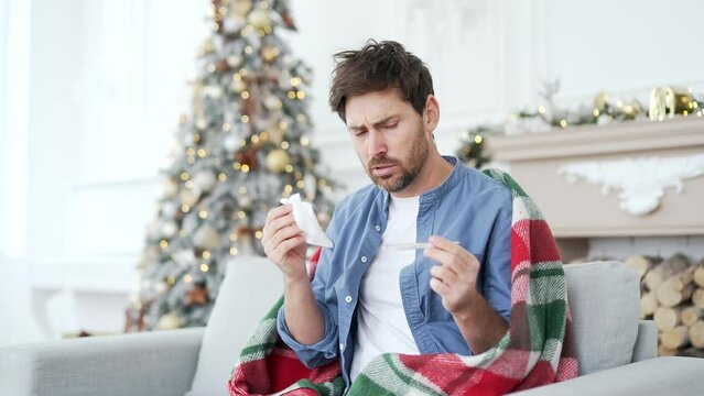 Sick man with a cold or flu wrapped in a blanket sitting on sofa at home during winter New Year Christmas Xmas holidays. Guy holds a thermometer in his hands, coughs, wipes his nose with handkerchief