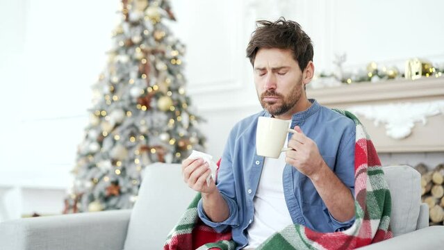 Sick man with a cold or flu is suffering from a cough sitting on sofa at home during winter New Year Christmas Xmas holidays. Male wrapped in blanket wipes his nose with a handkerchief, drinks hot tea