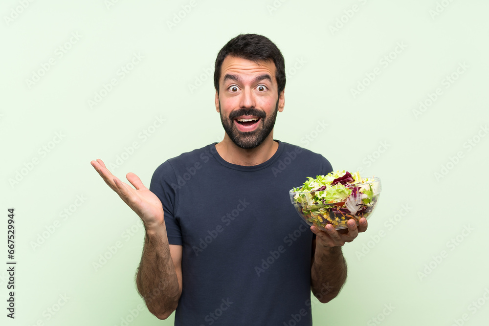 Wall mural young handsome man with salad over isolated green wall with shocked facial expression