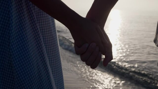 Couple holding hands near sea at sunset, closeup. Slow motion effect