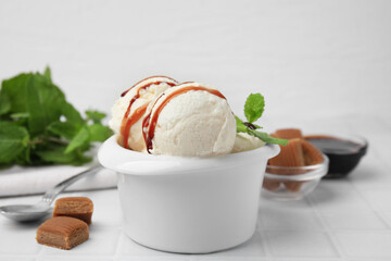 Scoops of tasty ice cream with mint leaves, candies and caramel sauce on white tiled table, closeup