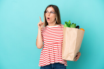 Young woman holding a grocery shopping bag isolated on blue background thinking an idea pointing...