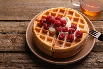 Tasty Belgian waffles with fresh raspberries, whipped cream, cup of tea and fork on wooden table, closeup