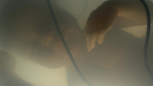Close up of silhouette of sleeping lab grown baby in late stage development floating in lab made amniotic fluid inside artificial womb and connected to nutrition system via wires