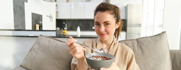 Close up portrait of cute woman on sofa, eating breakfast, holding delicious chocolate cereals with milk in a bowl, having proper morning meal