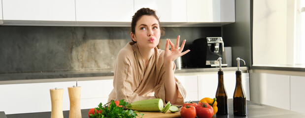 Portrait of beautiful young woman in bathrobe, cooking meal for family, shows chefs kiss, okay sign, making food, preparing vegetarian dinner, chopping vegetables