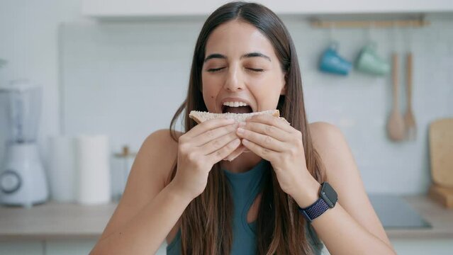 Video of  beautiful sporty woman eating healthy sanwich while looking at camera in the kitchen at home