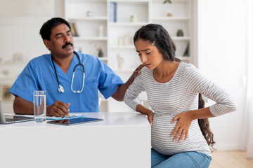 Indian Doctor Man Comforting Pregnant Female Feeling Unwell During Appointment In Clinic