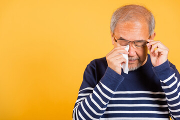 Portrait Asian senior old man sad wiping away his tears studio shot isolated on yellow background,...