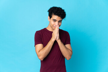 Young Venezuelan man isolated on blue background keeps palm together. Person asks for something