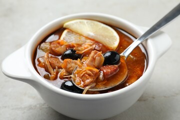 Meat solyanka soup with sausages, olives, vegetables and spoon in bowl on white textured table, closeup