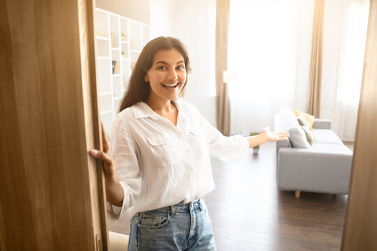 Cheerful young Indian woman inviting inside, standing by an open door