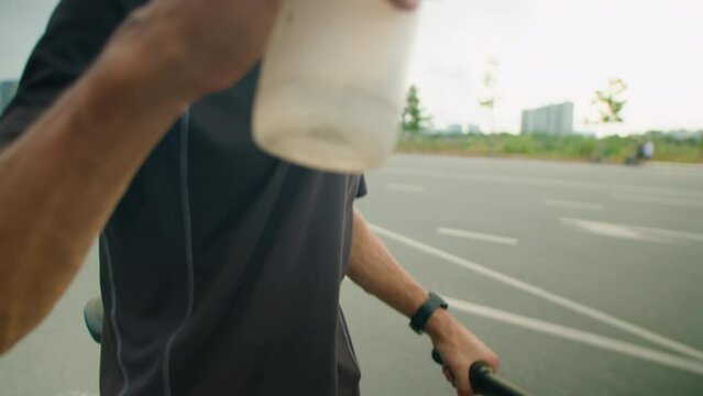 Tilt up shot of thirsty cyclist in riding clothing and helmet standing with bike on road and drinking water from bottle