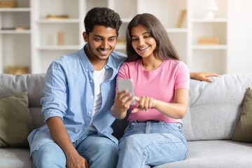 Obraz premium Happy Indian couple on sofa using smartphone, woman pointing at phone screen