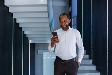 Mature successful boss in shirt walking down corridor of business building inside, man using phone,...