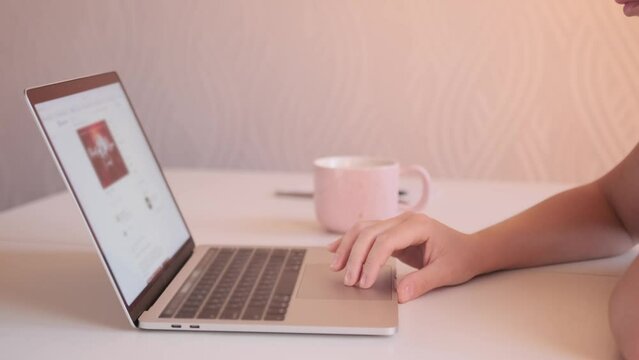 Side view of woman's hand scrolling social media feed on touchpad of laptop, close up, looking out for advertisements and photos on networks, checking morning mail next to cup of tea in kitchen