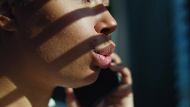 Side view of successful multicultural female talking on personal smartphone while standing near office window with jalousie. Busy woman with plump lips resolving work issues indoors.