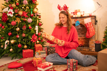 Obraz na płótnie Canvas Happy girl holding christmas gift box with smile hild wearing Chrismas jumper posing on floor near fireplace and xmas tree.