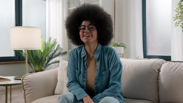 African American woman ethnic girl at home couch sofa looking at camera smiling laughing say wow amazed excited lady homeowner wonder surprised happy smile indoors in living room good news reaction