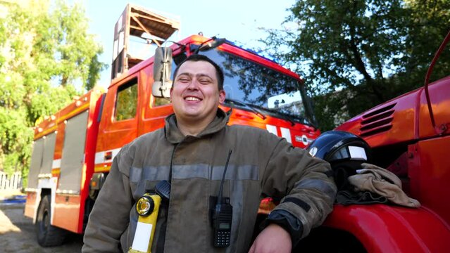 Portrait of happy young fireguard in uniform against background of a fire truck. Male firefighter looking into camera and smiling near big red car. Concept of saving lives and heroic profession