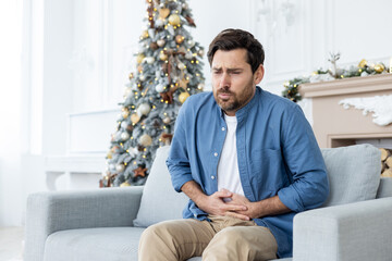 A young man suffers from severe abdominal pain during the Christmas holidays. He sits at home on the sofa near the Christmas tree and holds his body with his hands, grimacing from spasms - obrazy, fototapety, plakaty