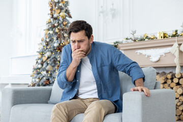 A young man sits at home on the couch during the New Year holidays and feels very nauseous, covers...