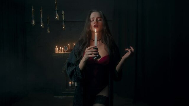 Young sexy brunette with a candle in her hands in a dark room. A young witch or vampire with a candle in her hands in a dark room. Divination by candle in a dark room