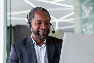 Mature experienced man in business suit with headset phone, working inside office with laptop,...