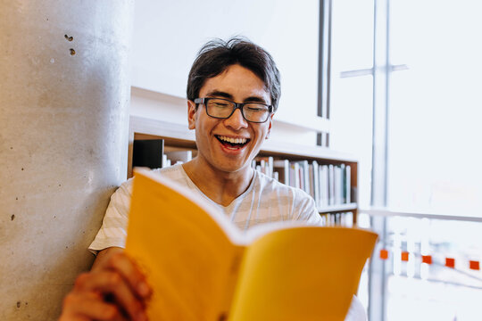 Portrait of a smiling man holding an opened yellow book with a bookshelf behind him.