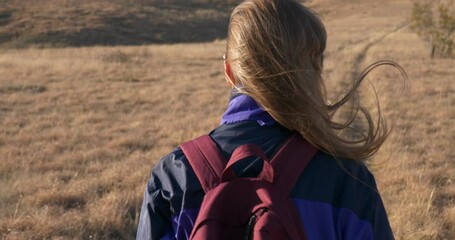 Rear close-up view of a young adult female hiker with a backpack walking along a trail on a hillside in the rays of an autumn sunset. Hair flutters in the wind. Slow motion. The camera follows