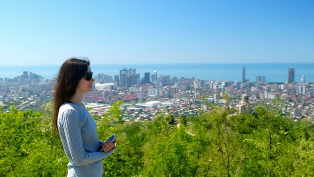 Woman tourist admire cityscape of Batumi city and sea from mountain in Georgia. Coastal town. Happy female in sunglasses smiling. Travel, tourism, wanderlust, adventure, explore, visiting attractions.