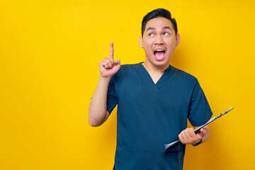 Excited professional young Asian male doctor or nurse wearing a blue uniform holding clipboard and pointing finger up, having great ideas isolated on yellow background. Healthcare medicine concept - obrazy, fototapety, plakaty
