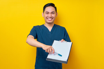 Professional young Asian male doctor or nurse wearing a blue uniform pointing with pen at blank paper on clipboard for patient signature approval isolated on yellow background