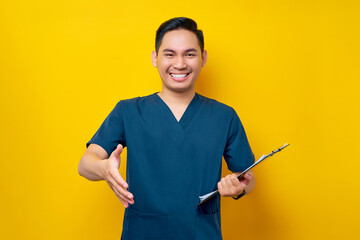 Smiling friendly professional young Asian male doctor or nurse wearing a blue uniform standing confidently holding a clipboard and greeting patients in a hospital isolated on yellow background