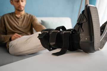 Young man with ankle orthosis, walker boot on leg sitting on the sofa at home, close-up.