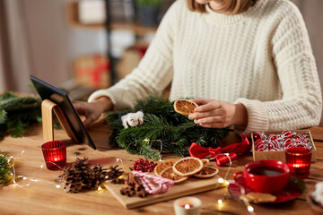 winter holidays, diy and hobby concept - close up of woman with tablet pc computer making christmas fir wreath at home - obrazy, fototapety, plakaty