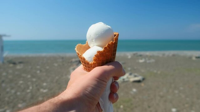 Hand of man holding ice cream on resort sea shore coast on blur background. Man resting relaxing on resort on vacation in sunny summer day. Travel, tourism, vacation, pleasure, enjoying.