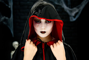 portrait of little boy as vampire in black and red hood with scary make up in dark studio on Halloween