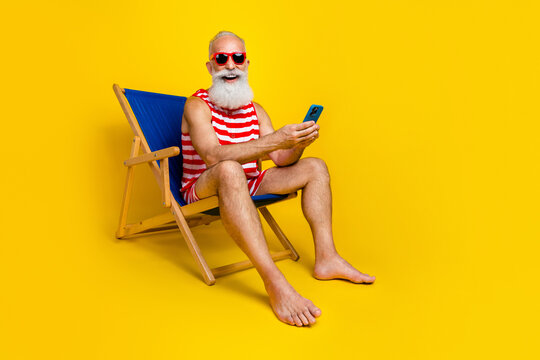 Full length photo of cheerful aged person sit lounger use smart phone have good mood isolated on yellow color background