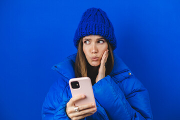 Shocked woman wearing casual blue hat and puffer jacket opens mouth hold phone reading advert...