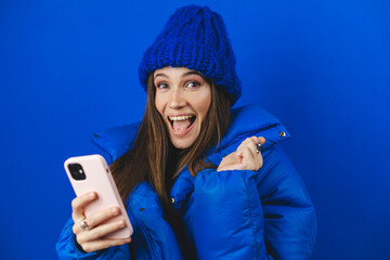 Fun woman hold in hand mobile cell phone doing winner gesture clenching fists isolated on blue background. Girl wear puffer, hat using smartphone apps winning online celebrating discount gift voucher.