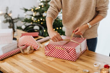 woman's hands wrapping and decorating a christmas gift with a christmas tree on the background in natural light