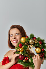 festive season, radiant woman with shimmery makeup holding Christmas wreath on grey backdrop