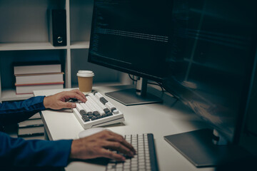 Male programmer working on desktop computer at white table in office, coding at night on simulated...