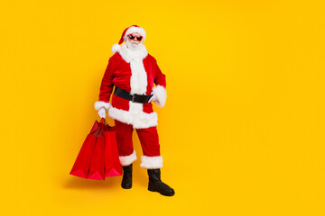 Full size photo of santa claus man wearing red costume celebrate happy new year holding shopping bags isolated on yellow color background
