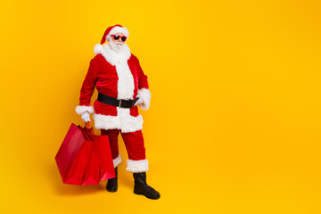 Fototapeta na wymiar Full length photo of cool santa claus hold shopping bags proposition for new year holiday preparation isolated on yellow color background