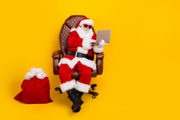 Full length portrait of eccentric santa sit chair use netbook new year presents bag empty space isolated on yellow color background